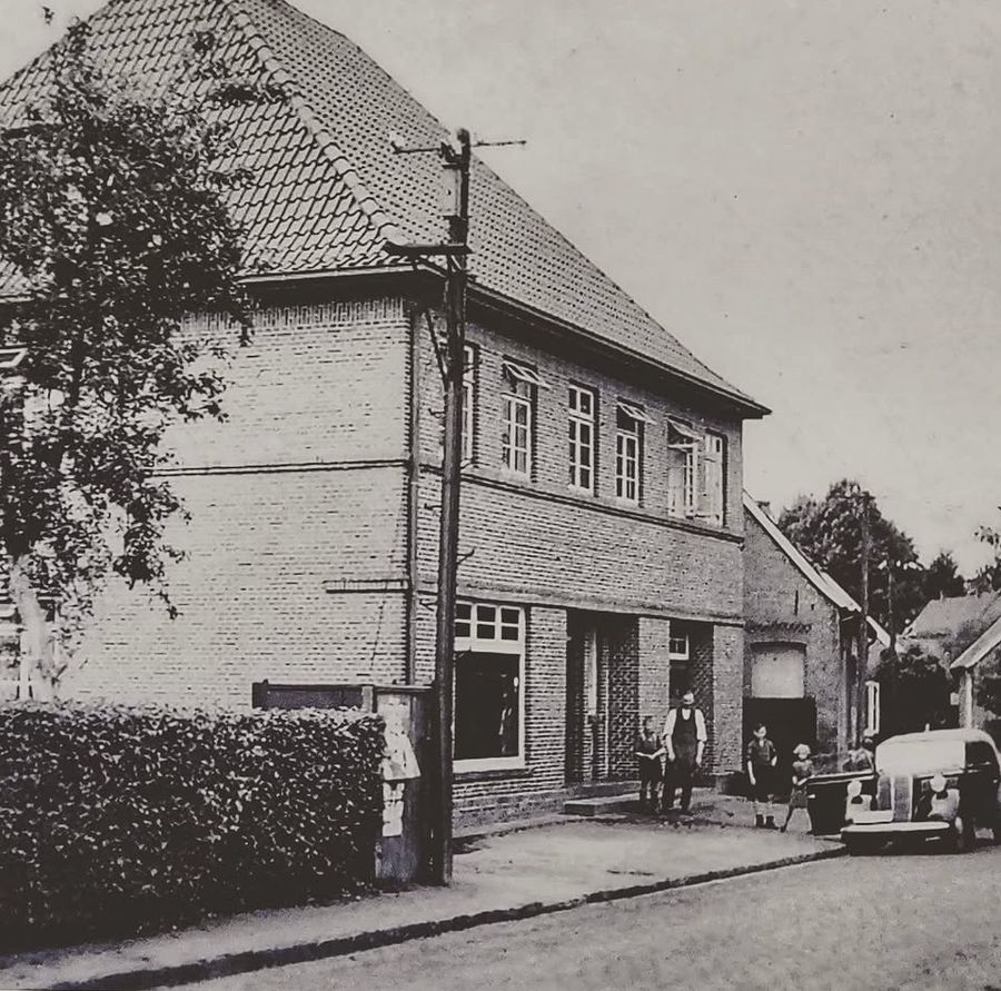 Schwarzweißfoto von 1954: ein zweigeschossiges Backsteinhaus an der Straße, davor mehrere Personen und ein Lieferwagen.
